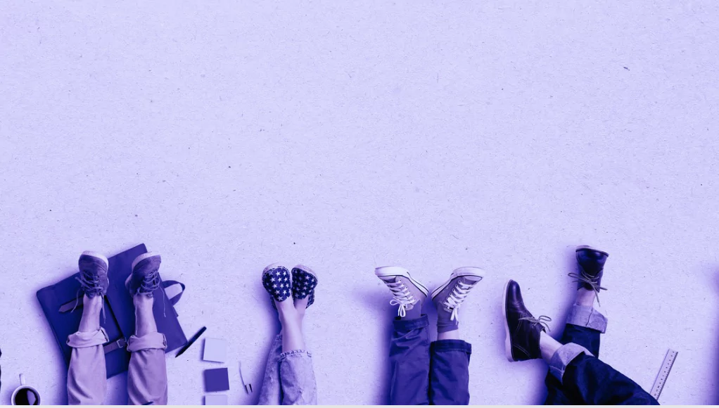 Vier Personen liegen entspannt nebeneinander auf dem Boden, nur ihre Beine und Schuhe sind sichtbar. Neben ihnen liegen Arbeitsutensilien wie Laptop, Notizbuch und Kaffeetasse.